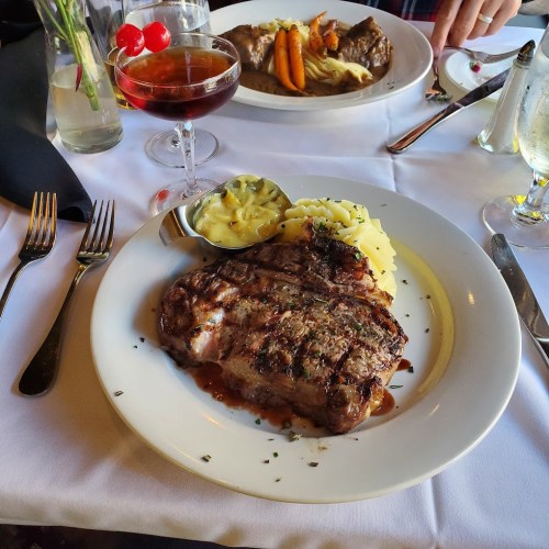 A grilled steak on a plate with mashed potatoes and a side of sauce, accompanied by a cocktail and another dish in the background.