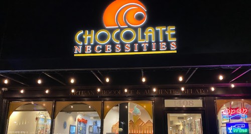 Storefront of "Chocolate Necessities" featuring a bright sign and open neon light, showcasing artisan chocolate and gelato.