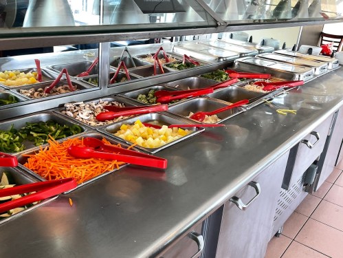 A salad bar with various fresh vegetables and fruits in metal trays, featuring red serving utensils.