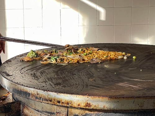 A chef stirs noodles and vegetables on a large, round griddle in a kitchen setting.