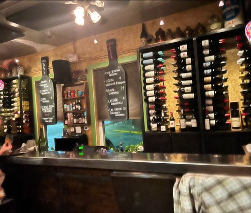 A cozy bar with a wooden counter, shelves lined with wine bottles, and a chalkboard menu in a warm, inviting atmosphere.