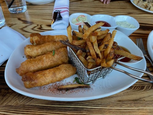 A plate of crispy spring rolls with a side of golden fries in a metal basket, accompanied by dipping sauces.