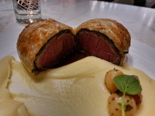 A plated dish featuring a sliced beef Wellington with pink center, creamy mashed potatoes, and garnished vegetables.