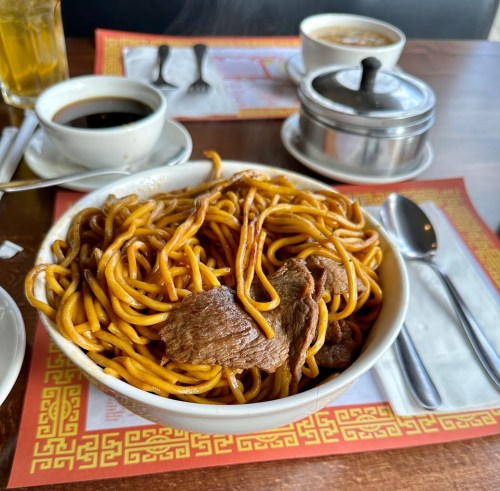 A bowl of beef lo mein noodles sits on a table with a cup of coffee and a glass of iced tea in the background.