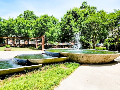 A modern fountain with three tiers, surrounded by lush greenery and trees under a clear blue sky.
