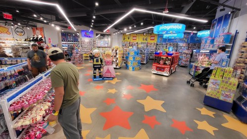 A colorful toy store interior with shelves of toys, a Playmobil display, and shoppers browsing among star-patterned flooring.