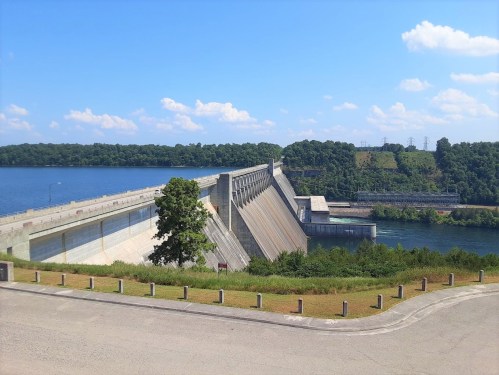 A large concrete dam spans a river, surrounded by greenery and under a clear blue sky with scattered clouds.