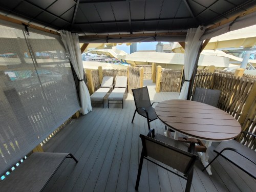 A cozy outdoor gazebo with a round table, chairs, and lounge chairs, surrounded by wooden fencing and a sunny sky.