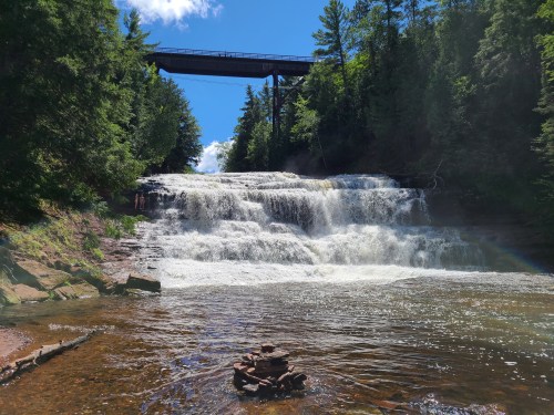 A scenic waterfall cascades down rocky steps, surrounded by lush greenery and a bridge overhead under a bright blue sky.