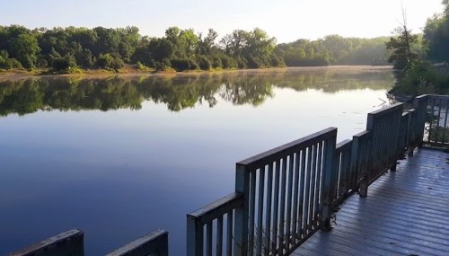 A calm river reflects trees and a clear sky, with a wooden deck in the foreground.