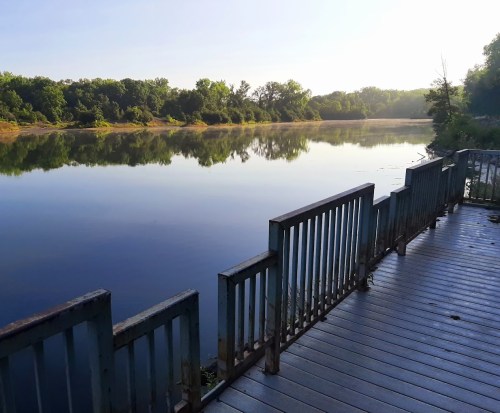 A serene river scene at dawn, with calm water reflecting trees and a wooden deck in the foreground.
