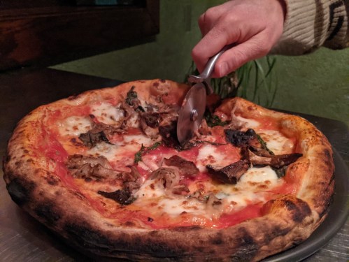 A hand using a pizza cutter to slice a freshly baked pizza topped with cheese and mushrooms.