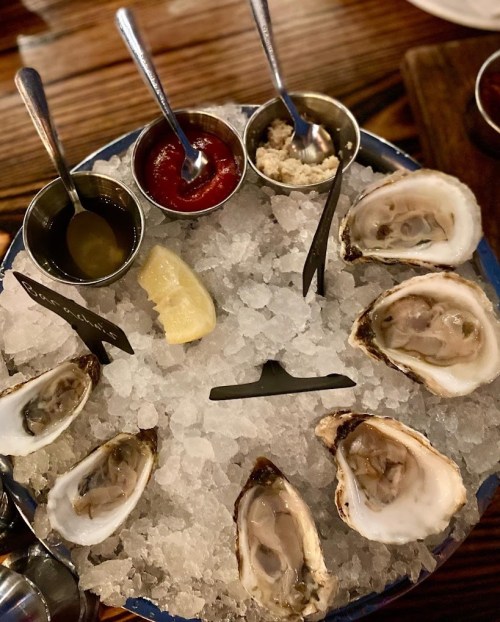 A platter of fresh oysters on ice, accompanied by sauces and a lemon wedge.