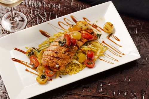 Grilled salmon on a bed of couscous and vegetables, garnished with cherry tomatoes and a drizzle of sauce.