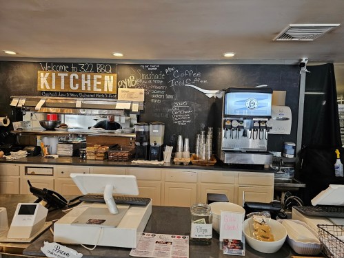Interior of a BBQ kitchen with a counter, menu board, coffee machine, and various condiments and utensils displayed.
