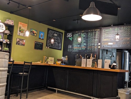 A cozy bar with a green wall, chalkboard menus, and a wooden counter with beer taps and seating.