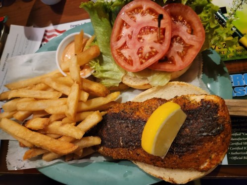 A plate with a seasoned fish fillet, lemon wedge, lettuce, tomato, and a side of crispy French fries.