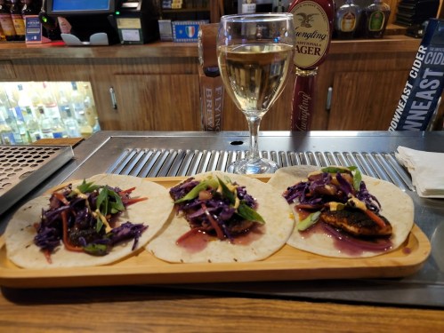 Three tacos on a wooden platter, topped with purple cabbage and vegetables, alongside a glass of white wine.
