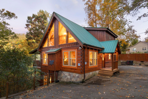 A cozy wooden cabin with a green metal roof, surrounded by trees and autumn foliage. Warm lights glow from the windows.