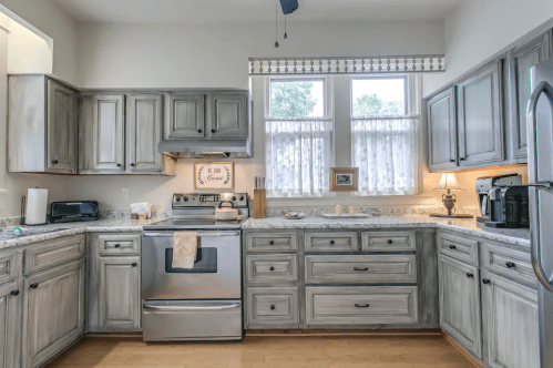 A modern kitchen with gray cabinets, stainless steel appliances, and a light countertop, featuring natural light from windows.