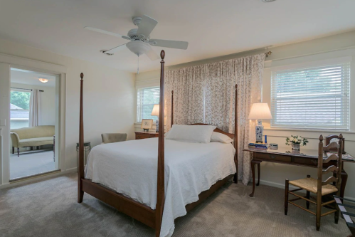 A cozy bedroom featuring a four-poster bed, a desk, and soft lighting, with a window and light-colored decor.