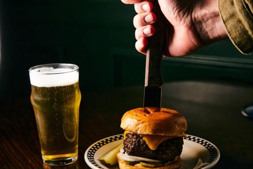 A hand holding a knife above a cheeseburger on a plate, with a glass of beer nearby.