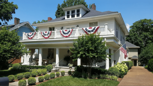 A charming two-story house adorned with red, white, and blue bunting, surrounded by well-maintained greenery and an American flag.