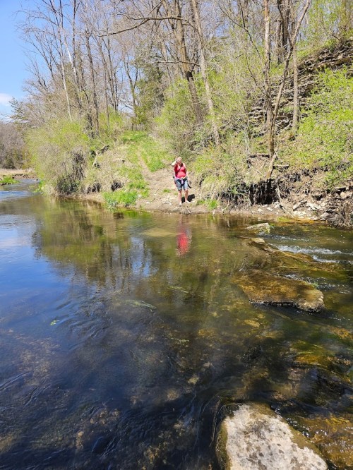 A person in a red jacket stands by a calm river, surrounded by trees and greenery, reflecting in the water.