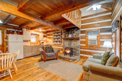 Cozy log cabin interior with a stone fireplace, wooden beams, and a kitchen area, featuring comfortable seating and rustic decor.