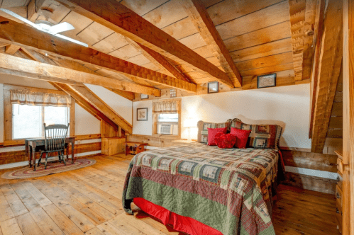 Cozy attic bedroom with wooden beams, a quilted bed, a small table, and a window letting in natural light.