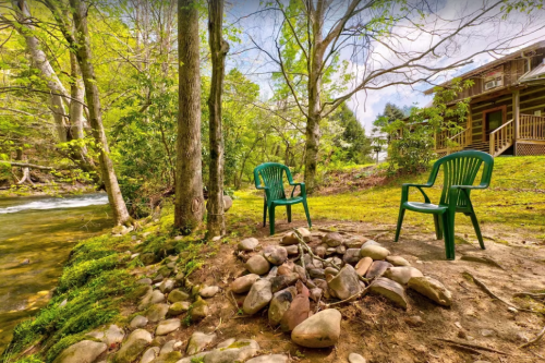 Two green chairs by a stone fire pit near a stream, surrounded by lush greenery and trees. A cabin is visible in the background.