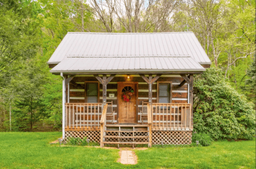 A charming log cabin with a front porch, surrounded by greenery and trees, featuring a wreath on the door.