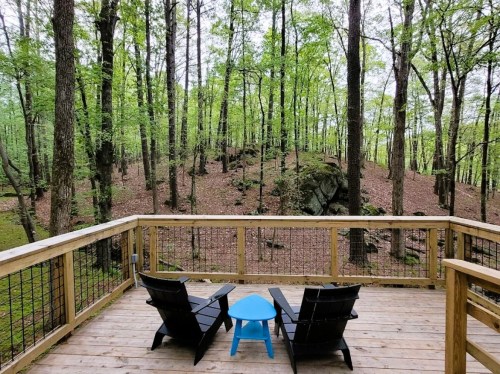 A wooden deck with two black chairs and a blue table, surrounded by lush green trees and rocky terrain.