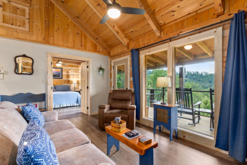 Cozy cabin interior with wooden walls, a sofa, armchair, and large windows overlooking a green landscape.