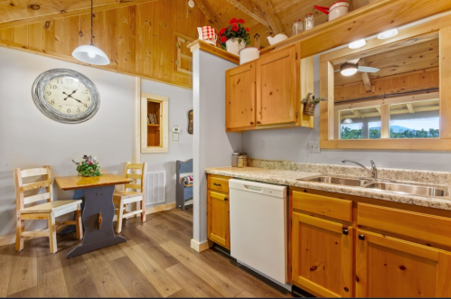 Cozy kitchen with wooden cabinets, a clock, a small dining table, and a window overlooking greenery.