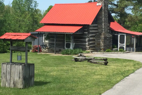 A rustic log cabin with a red metal roof, surrounded by green grass and trees, featuring a stone chimney and a nearby well.