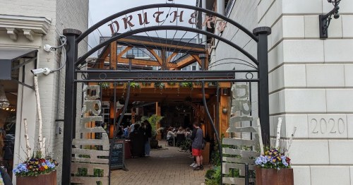Entrance to a lively restaurant with outdoor seating, surrounded by greenery and flowers.