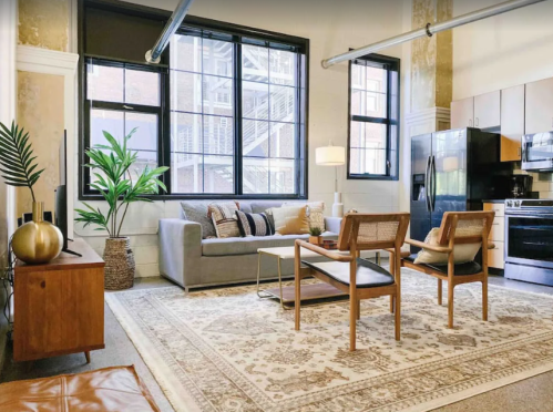 Cozy living room with a gray sofa, wooden chairs, a coffee table, and large windows, featuring a plant and decorative rug.
