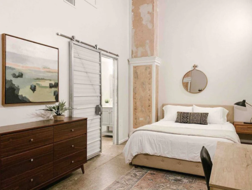 Cozy bedroom with a large bed, wooden dresser, round mirror, and sliding door, featuring soft colors and natural light.
