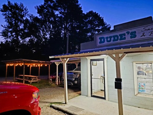 A small building labeled "Dude's" with outdoor seating, illuminated by string lights, at dusk. A red vehicle is parked nearby.