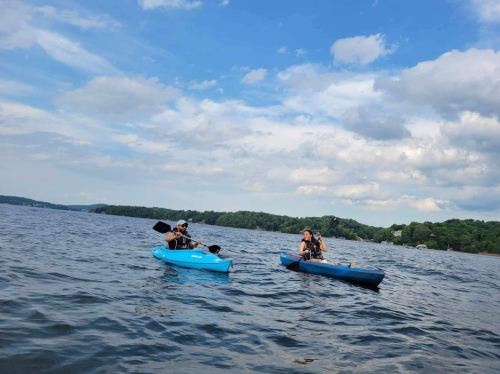 Two people kayaking on a calm lake under a partly cloudy sky, with lush greenery in the background.