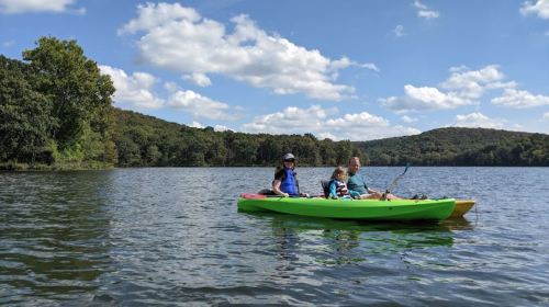 A family kayaking on a calm lake surrounded by green hills and a blue sky with fluffy clouds.