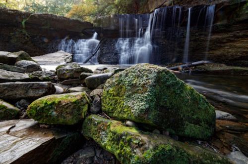 A serene waterfall cascades over rocks, surrounded by lush greenery and moss-covered stones in a tranquil forest setting.