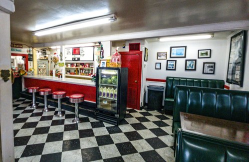A retro diner interior with checkered floors, green booths, red stools, and a counter displaying drinks and snacks.