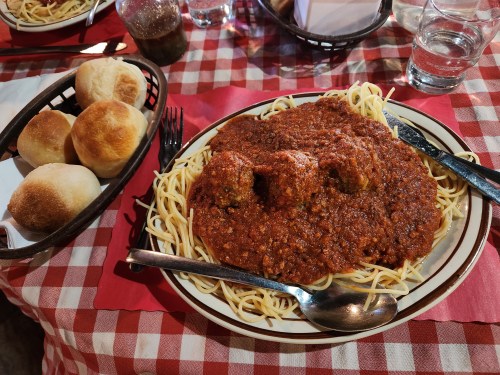 A plate of spaghetti topped with meatballs and marinara sauce, accompanied by a basket of rolls on a checkered tablecloth.