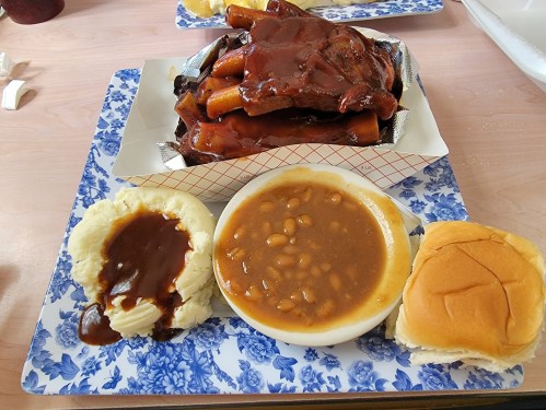 A tray with BBQ ribs, mashed potatoes with gravy, baked beans, and a soft roll on a floral-patterned plate.