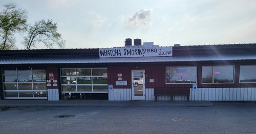 Exterior of a BBQ restaurant with a sign reading "Whatcha Smokin'? BBQ & Brew" and large windows.