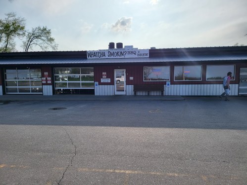 Exterior of "Whatcha Smoking' BBQ Brew" restaurant with a sign, large windows, and a person walking by.