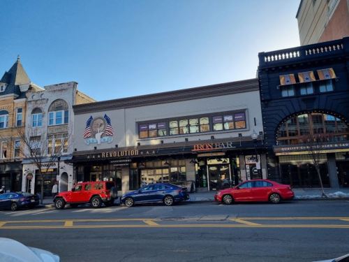 A street view of buildings featuring Revolution and Iron Bar, with parked cars in front and a clear blue sky above.