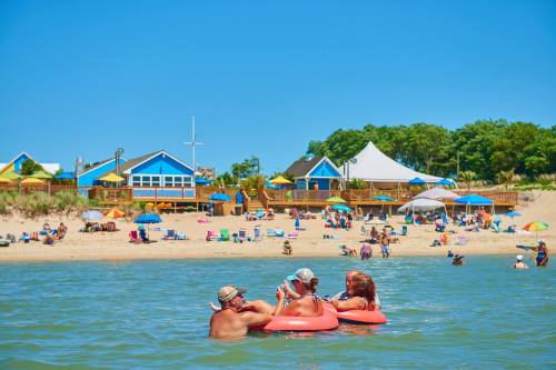 Three people in inflatable rings enjoy the water at a beach with colorful umbrellas and a lively crowd in the background.